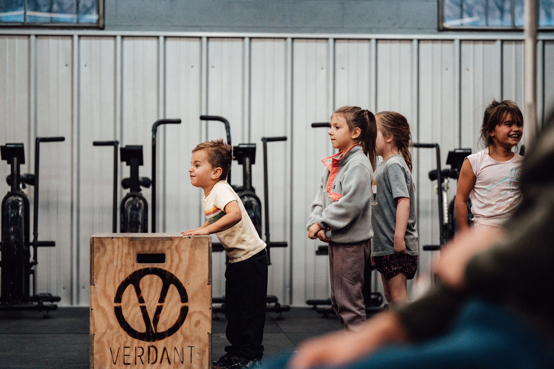 Children line up in gym, one on a wooden box, ready to jump. Others watch, anticipating the action.