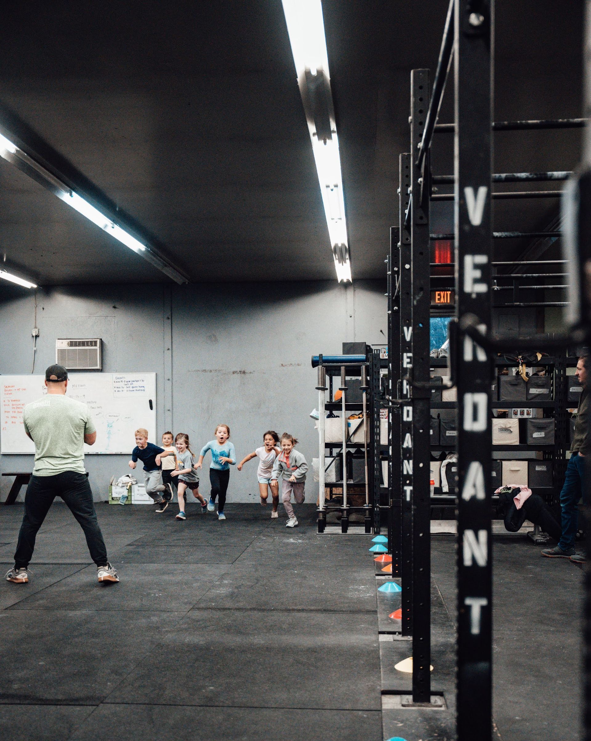 Children running towards a coach in a gym with black exercise racks.
