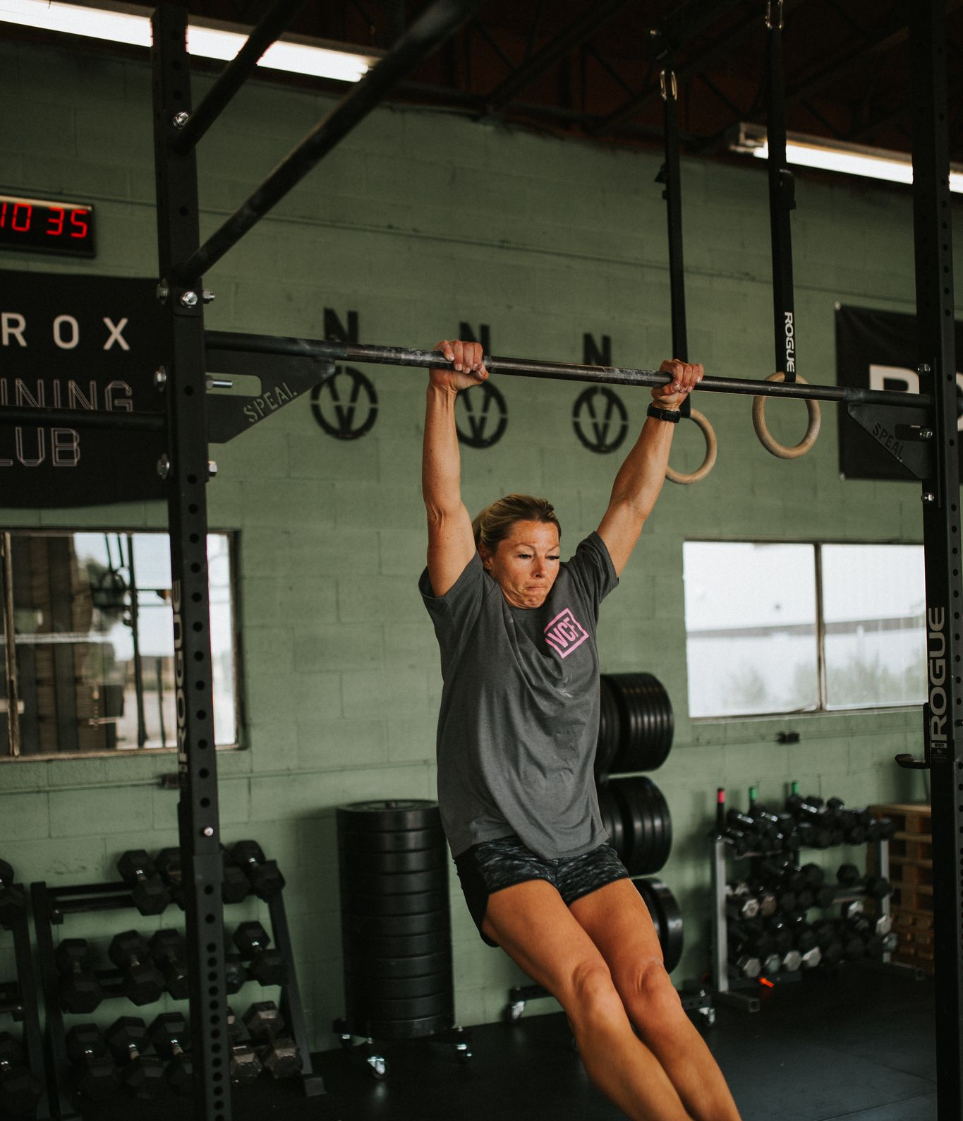 Woman hanging from a pull-up bar in a gym in Star, ID; arms extended, gray shirt, black shorts.