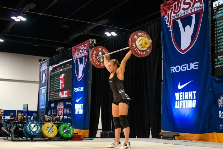 Female weightlifter in black lifting a barbell overhead at a USA Weightlifting competition.