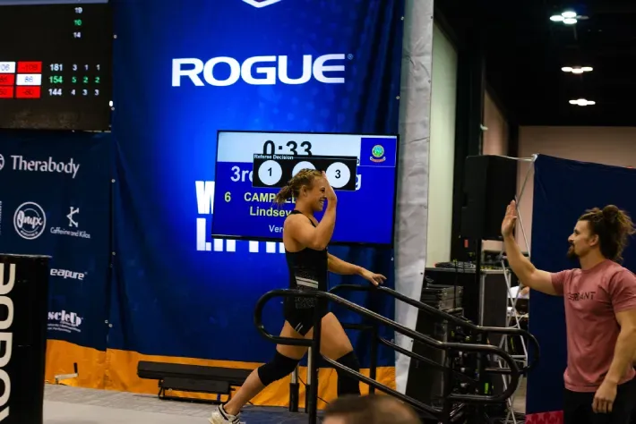 Female athlete high-fives coach after completing a stair step challenge at a Rogue competition. Blue, black setting.