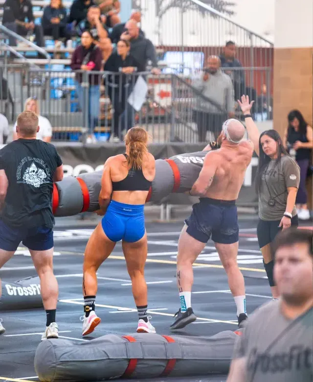 Athletes lifting heavy sandbags in a CrossFit competition. Spectators watch in a gym.