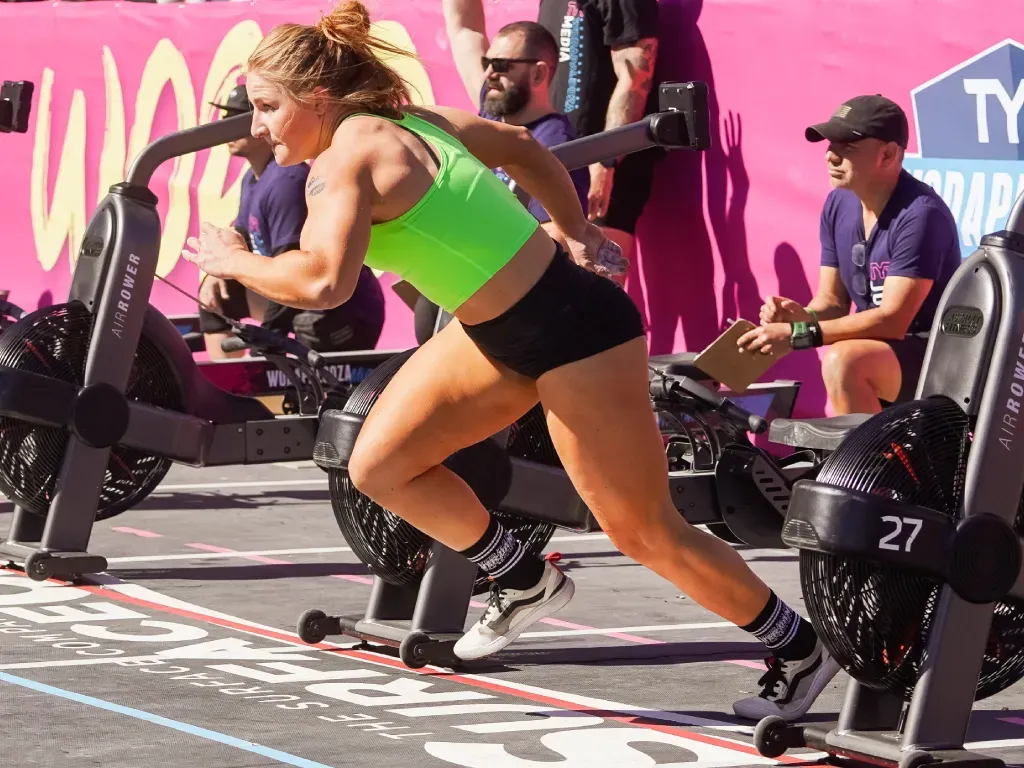 Female athlete running, likely in a competition, wearing a green top and black shorts; near air bikes.
