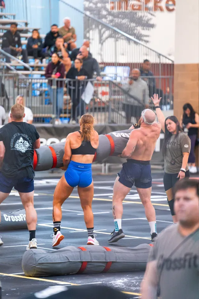 CrossFit athletes lifting heavy logs during a competition. Spectators watch in the background.