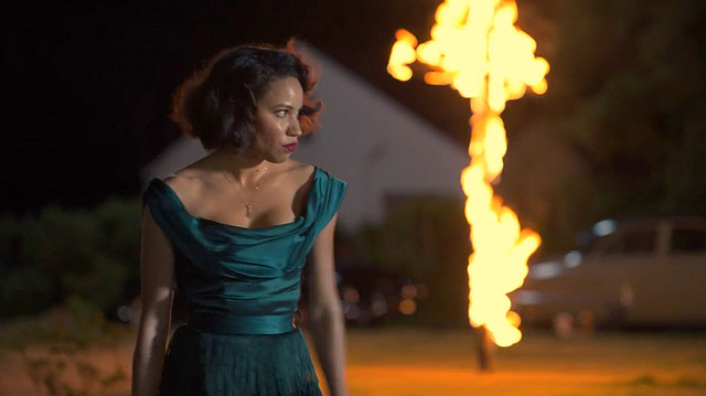 A black woman in a dark green dress looks angrily away from a burning cross a night. Leti Lewis, Lovecraft Country