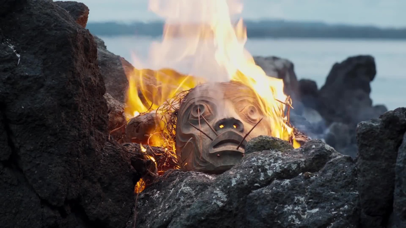 A Haida tribal mask burns on rocks by the seaside