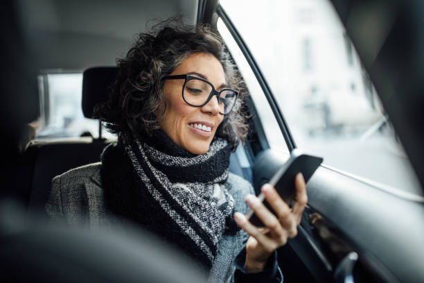 Woman wearing glasses and a scarf in a car, looking at a phone, smiling.