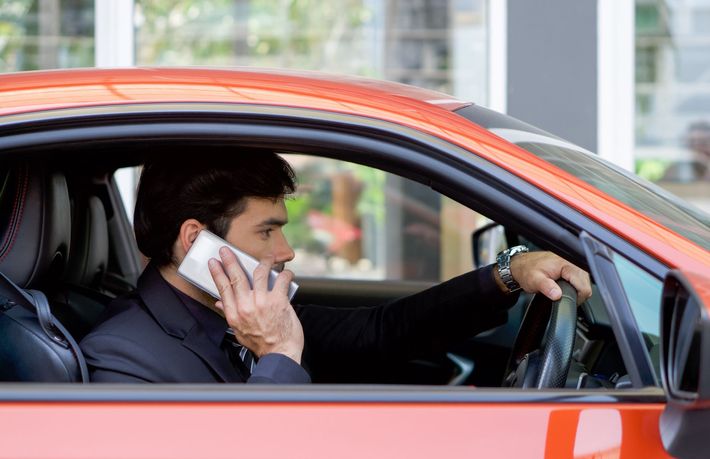 A man in a suit talks on his cell phone while driving an orange car.