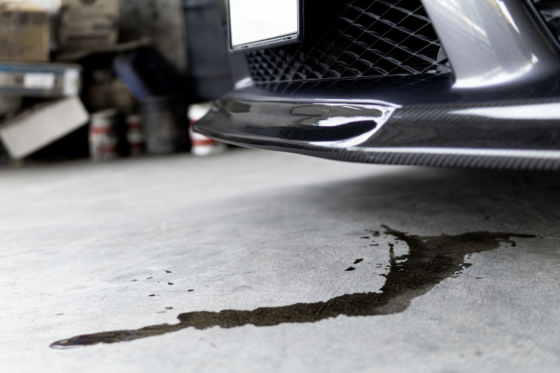 Black car's front end dripping a dark fluid onto a concrete floor, likely oil or another automotive fluid in a garage.