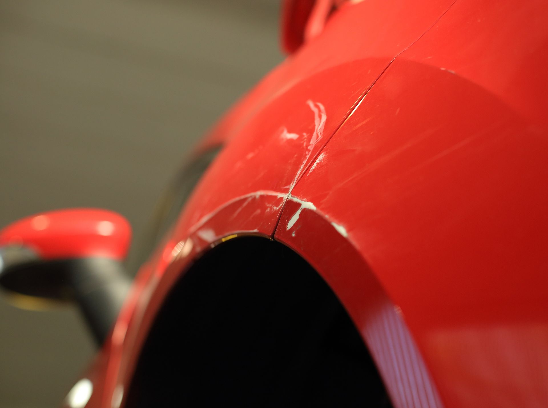 Close-up of a red car's rear quarter panel with white scratches and damage near the wheel arch.