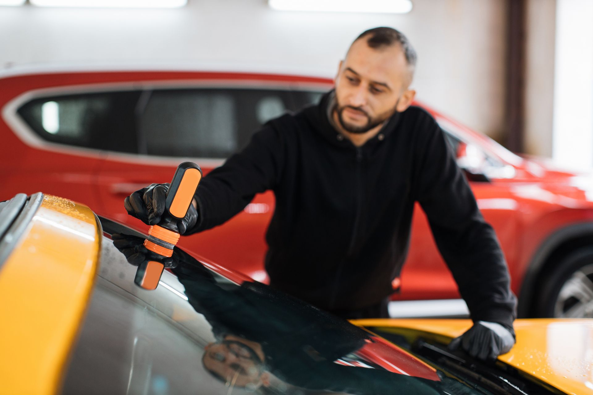 Man in black clothing repairing windshield of a yellow car in a garage, with a red car in the background.