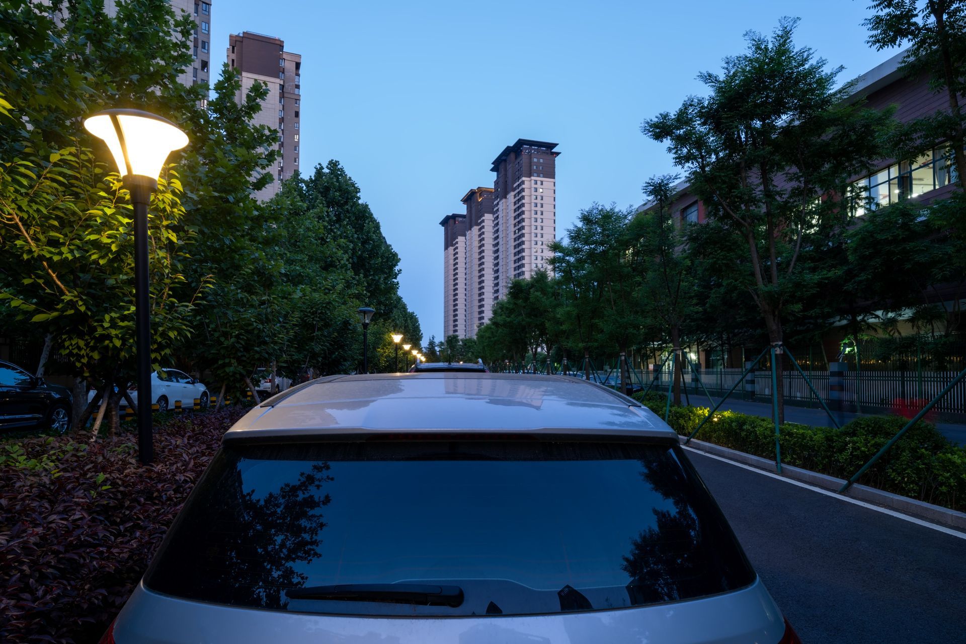 A silver car parked on a street lined with trees and lampposts, with apartment buildings in the background at dusk.