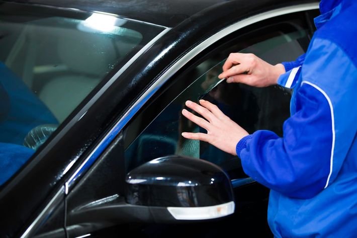 A person in a blue jumpsuit is applying a dark plastic visor to the side window of a black car.