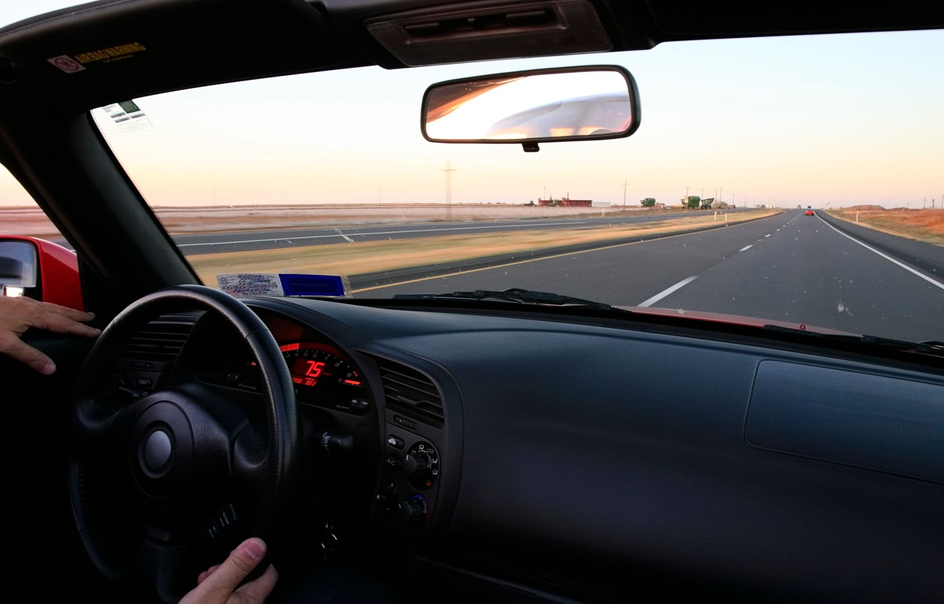 Interior view of a red convertible driving on an open road. Driver's hands on the steering wheel, with a dashboard and windshield in view.