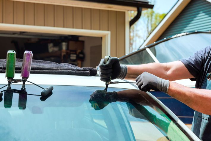 A person replacing a car windshield outdoors. They wear gloves and use a tool to remove the old glass.