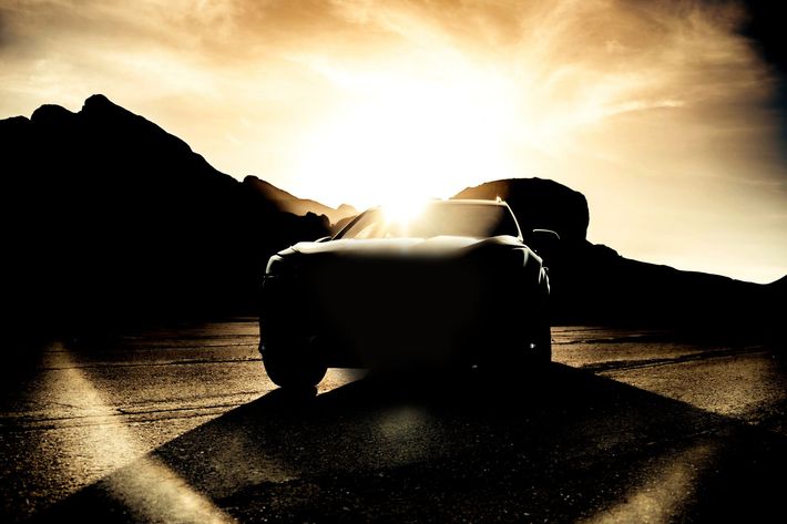 Silhouette of a vehicle driving toward the bright sun in a desert landscape, mountains in the background.