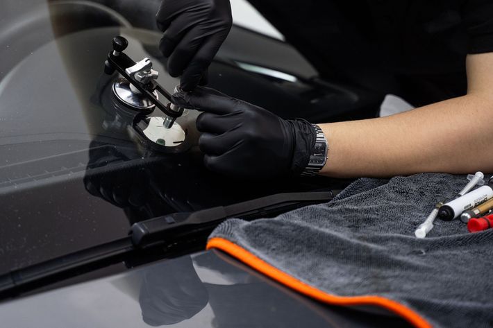 A person in black gloves repairing a windshield crack with a specialized tool on a car. Small tubes and a towel are nearby.