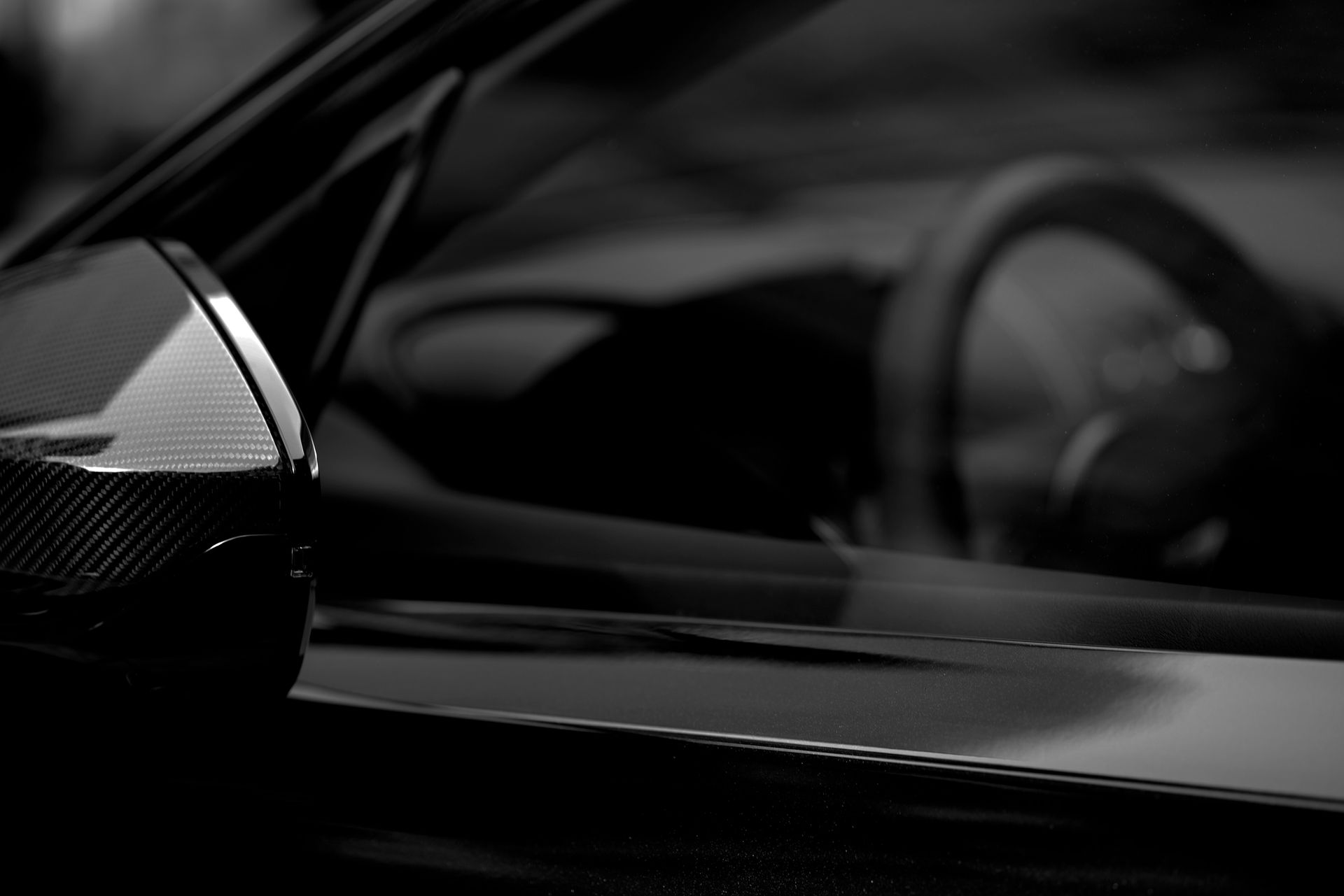 Black and white image of a car's side mirror with the interior visible. Shows the steering wheel, blurred reflections, and a sleek, modern design.