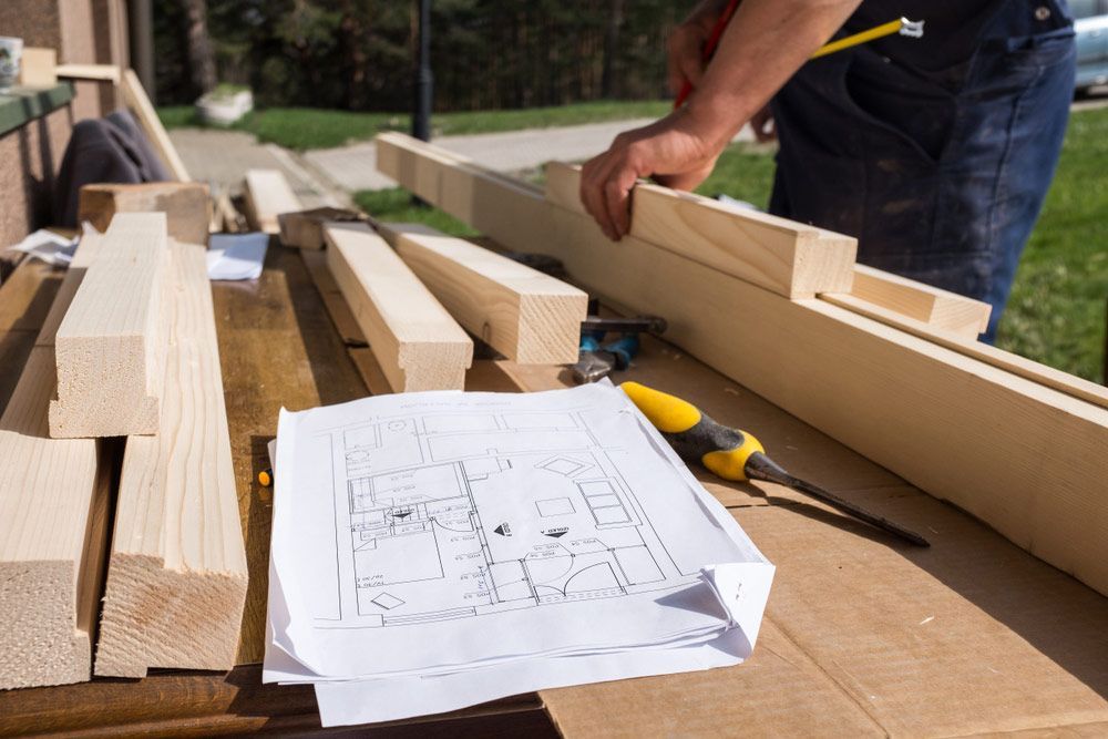 Person Measuring Wood With a Pencil Next to Blueprints and Lumber on a Table — CJ Custom Carpentry in Port Macquarie, NSW