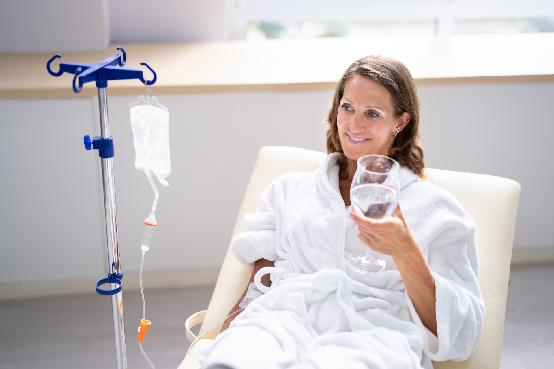 woman holding a glass full of water, lying down on a hospital bed with an iv beside her