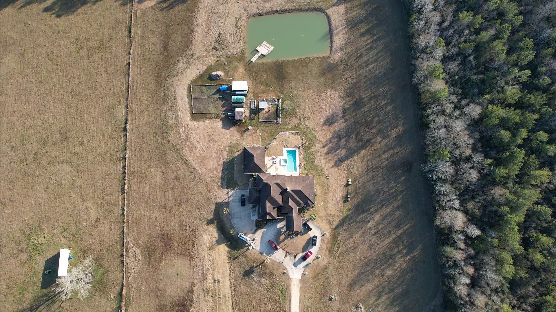 Aerial view of a house with a pool, pond, and small structures on a grassy property surrounded by forest.