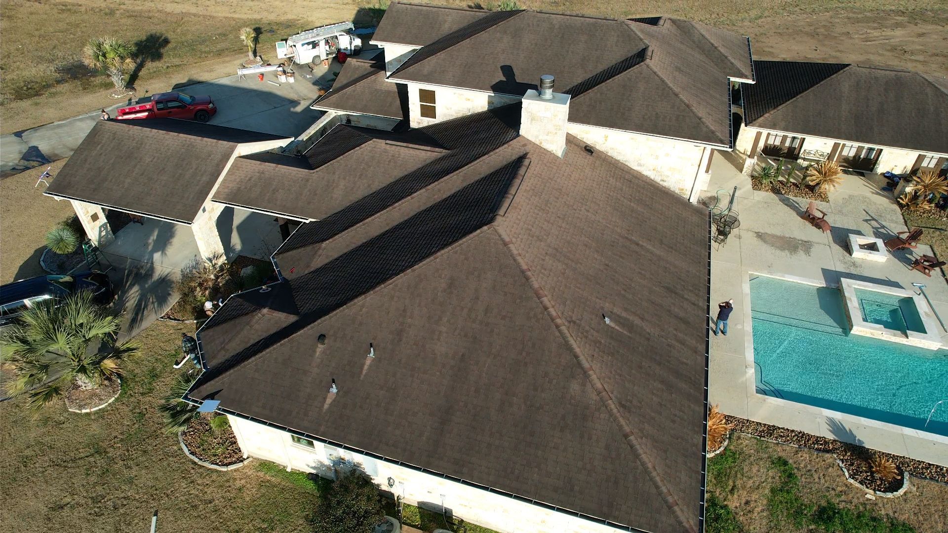 Aerial view of a large, brown-roofed house with a pool. The house is surrounded by a yard and dry landscape.