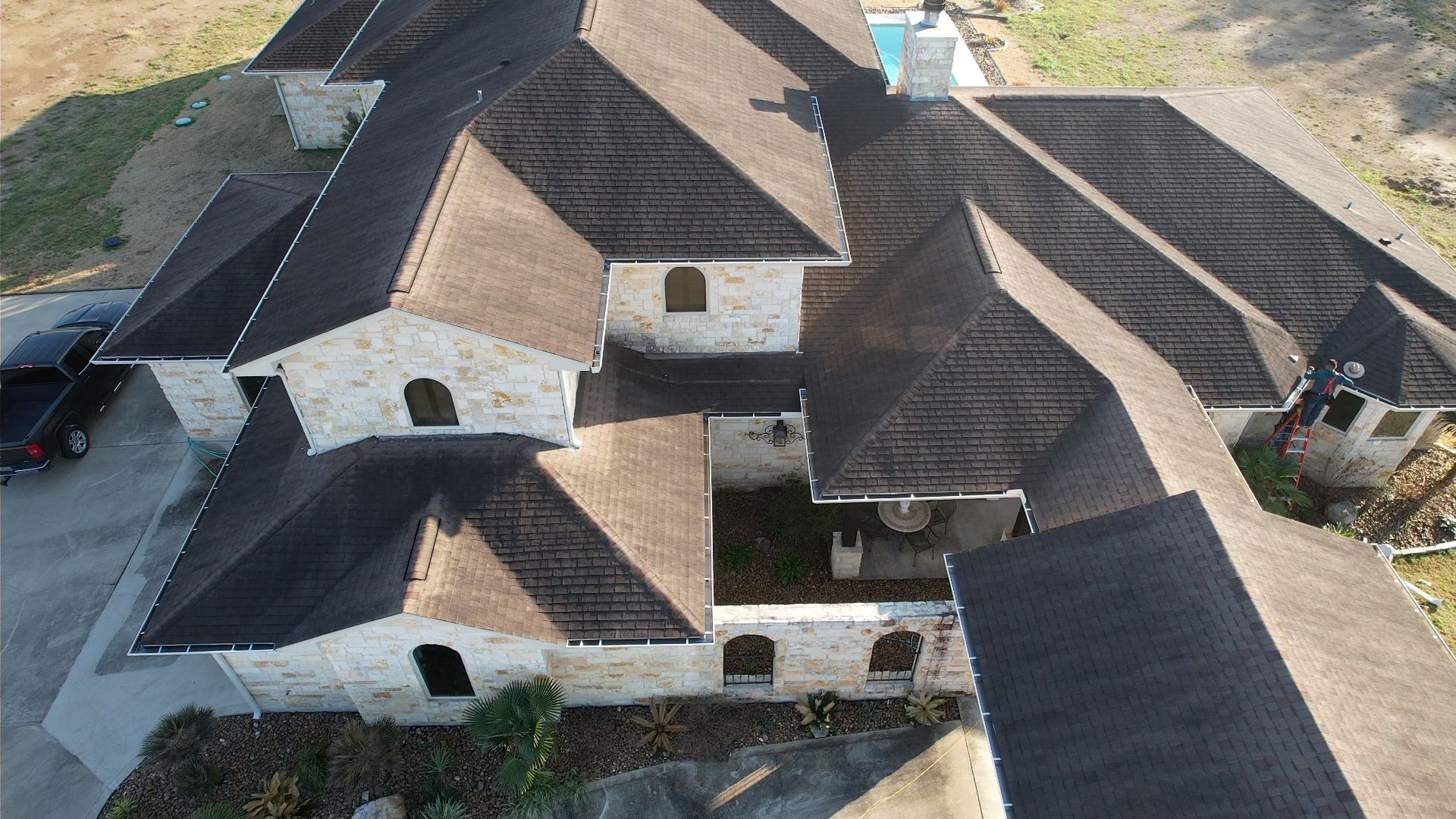 Aerial view of a large, multi-sectioned house with a dark roof and light-colored stone walls.