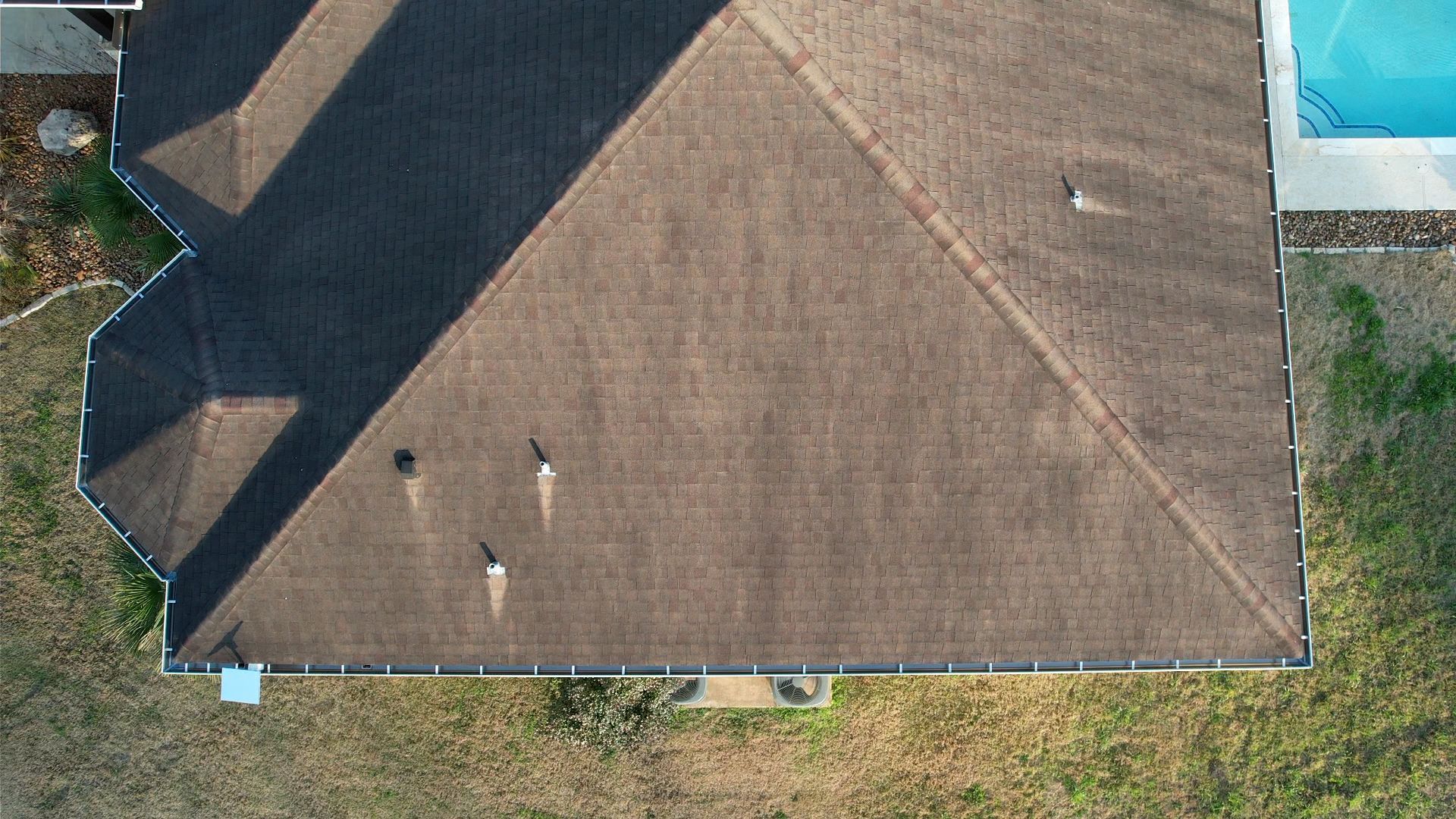 Overhead view of a brown asphalt shingle roof with several vents, near a swimming pool and grassy yard.