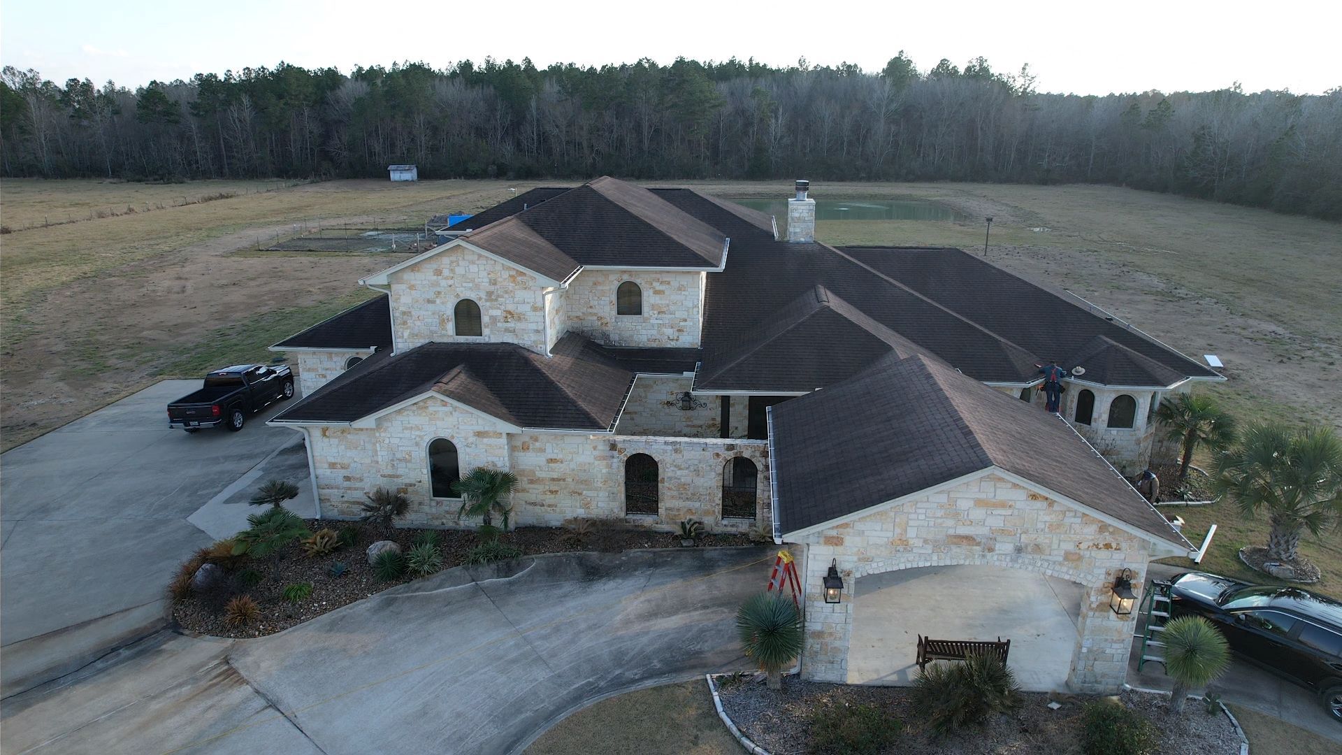 Stone house with dark roof and long driveway.  Truck and car parked outside.  Field and trees in background.