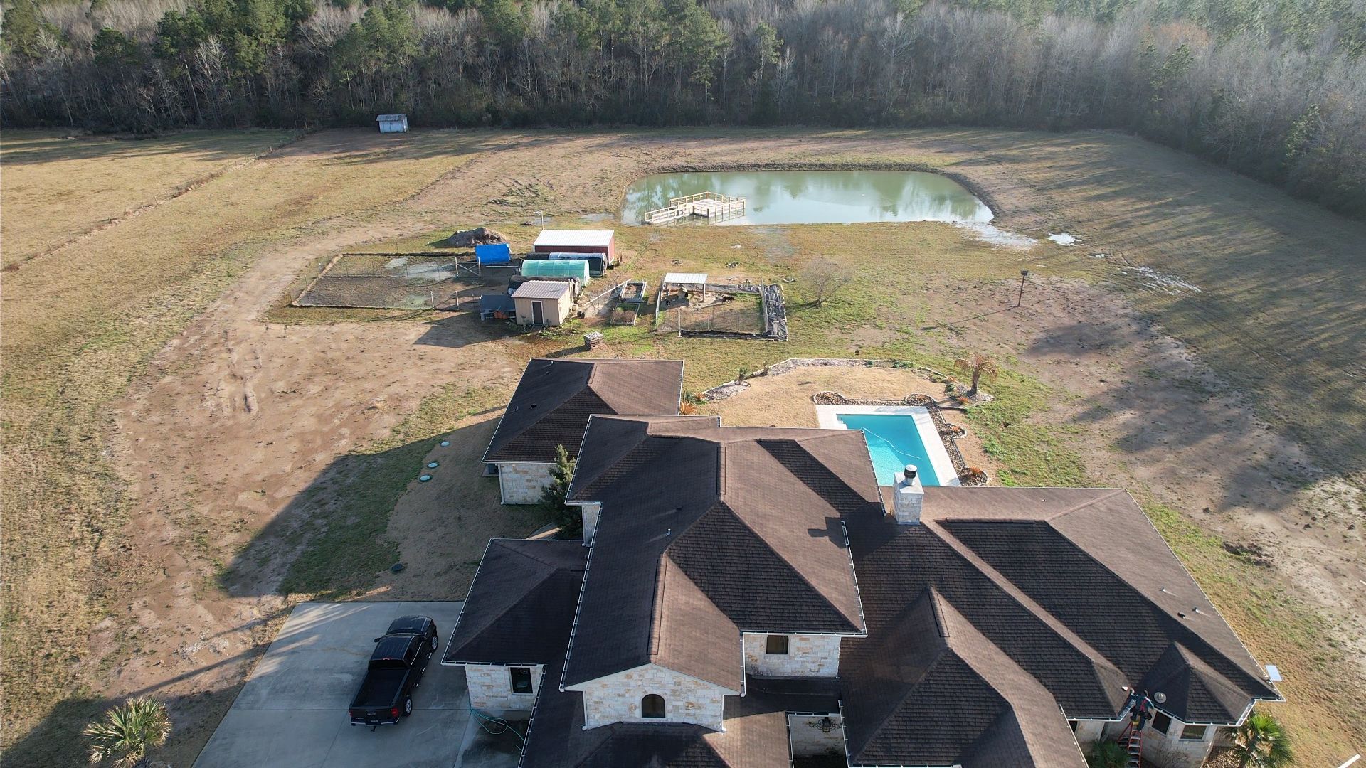 Aerial view of a house with a pool, pond, and outbuildings in a rural setting.