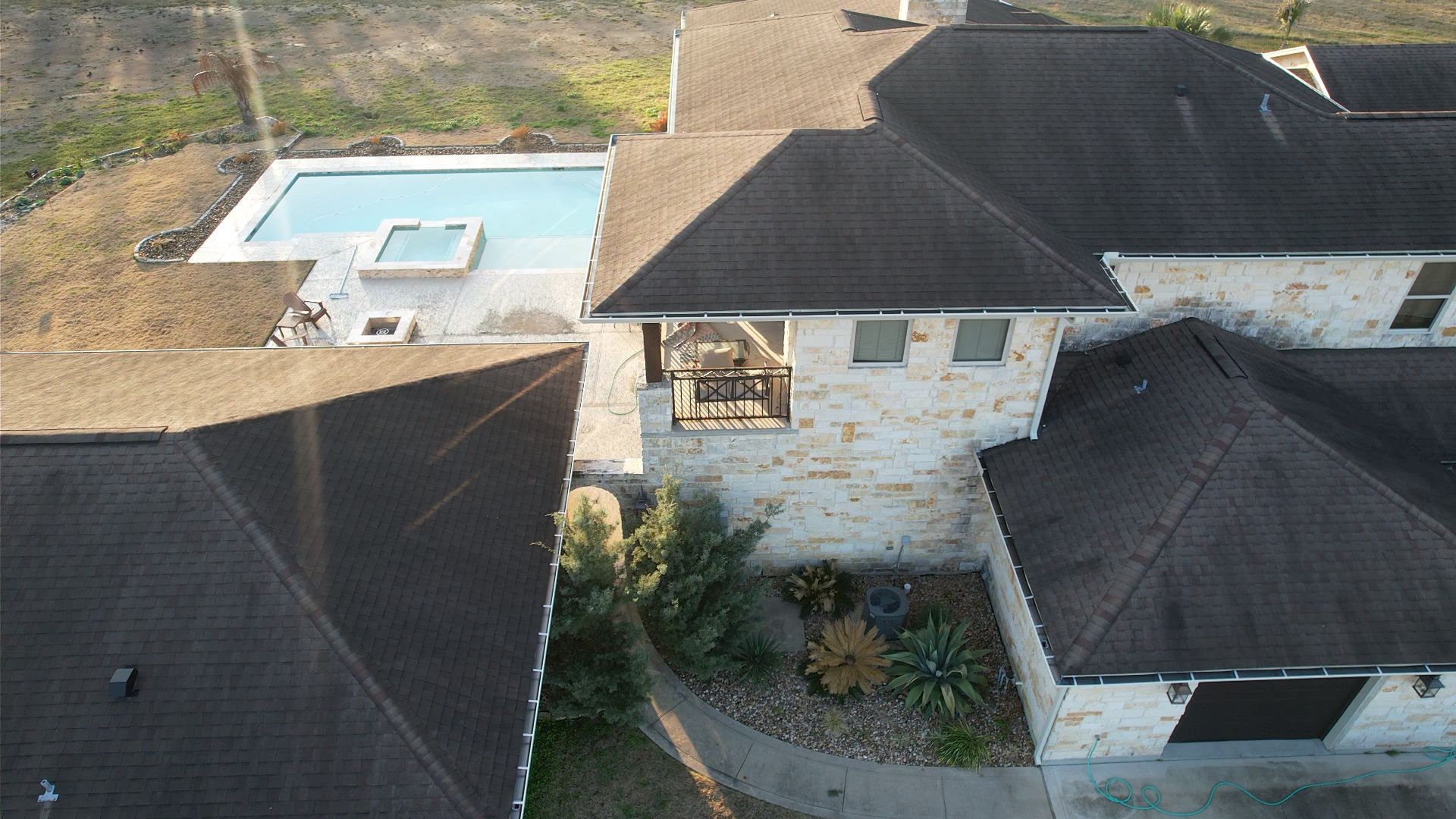 Drone view of a large house with a pool and dark roof; stone exterior; blue pool water.