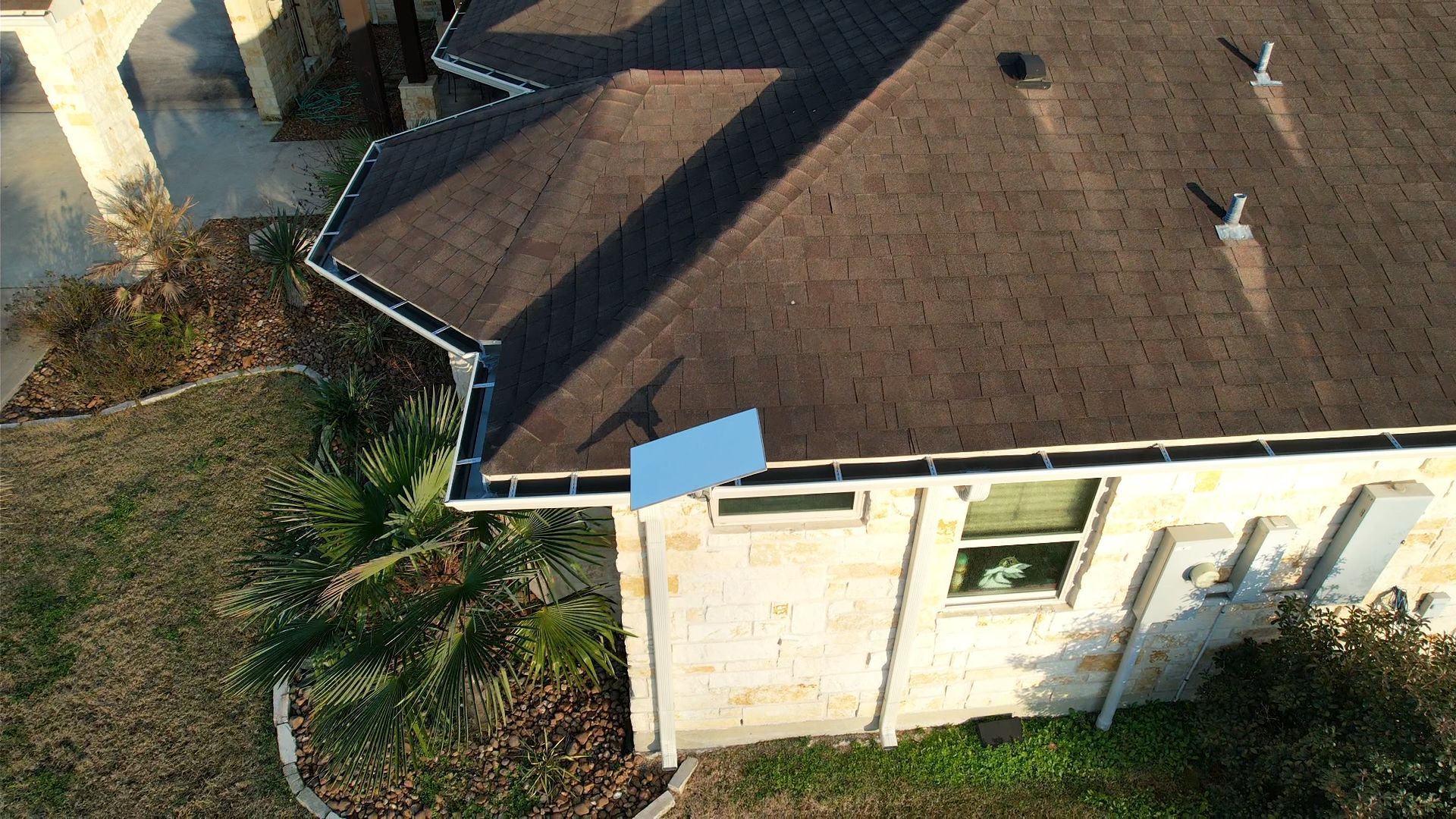 Overhead view of a house with a brown shingled roof, light-colored brick exterior, and a blue object on the roof.
