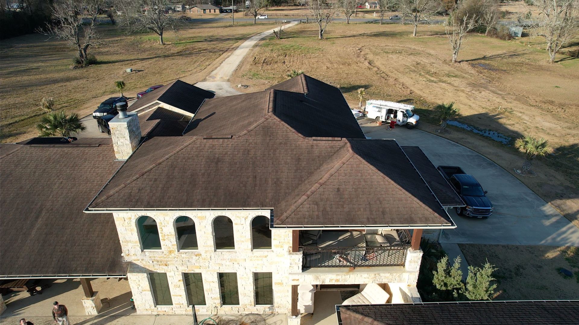 Aerial view of a large house with a brown roof and stone facade, driveway, and surrounding brown landscape.
