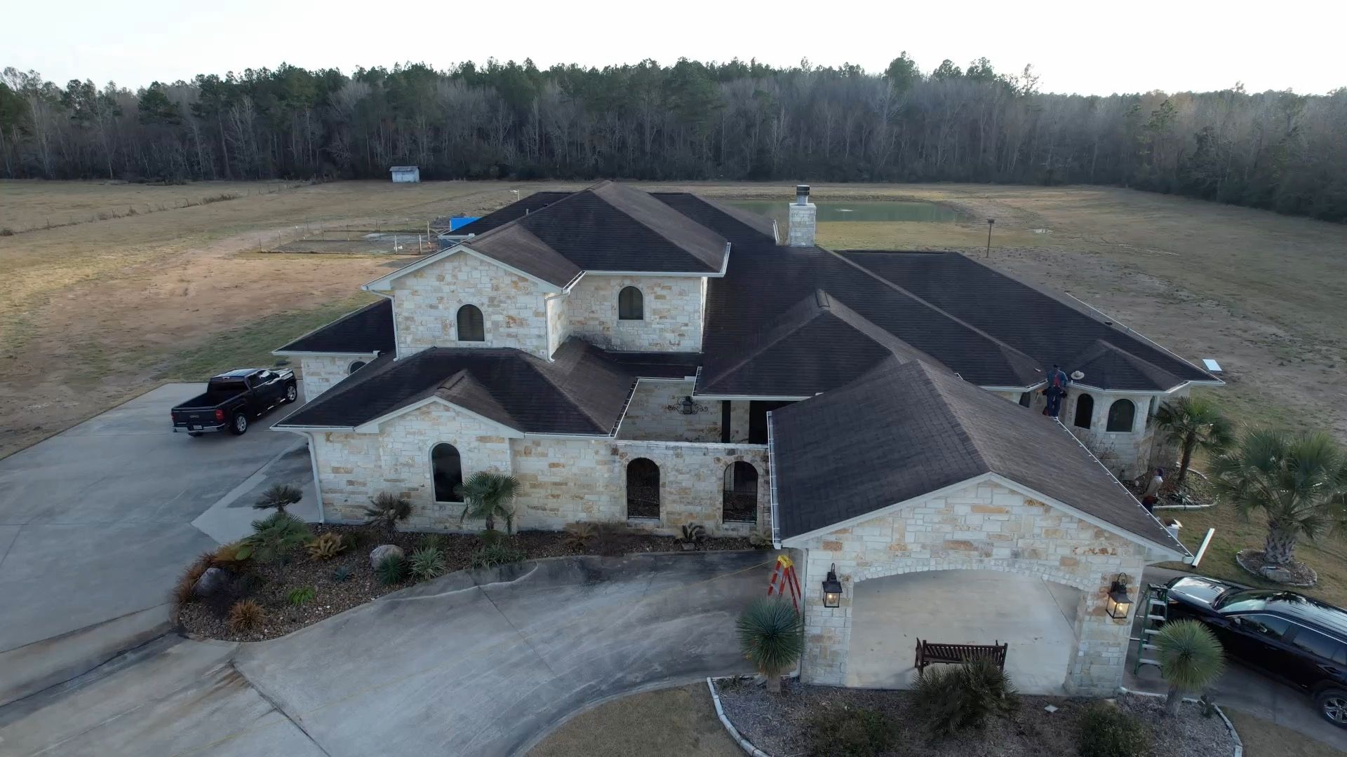 Large stone house with dark roof; driveway leads to the house, parked vehicles, and a field in the background.