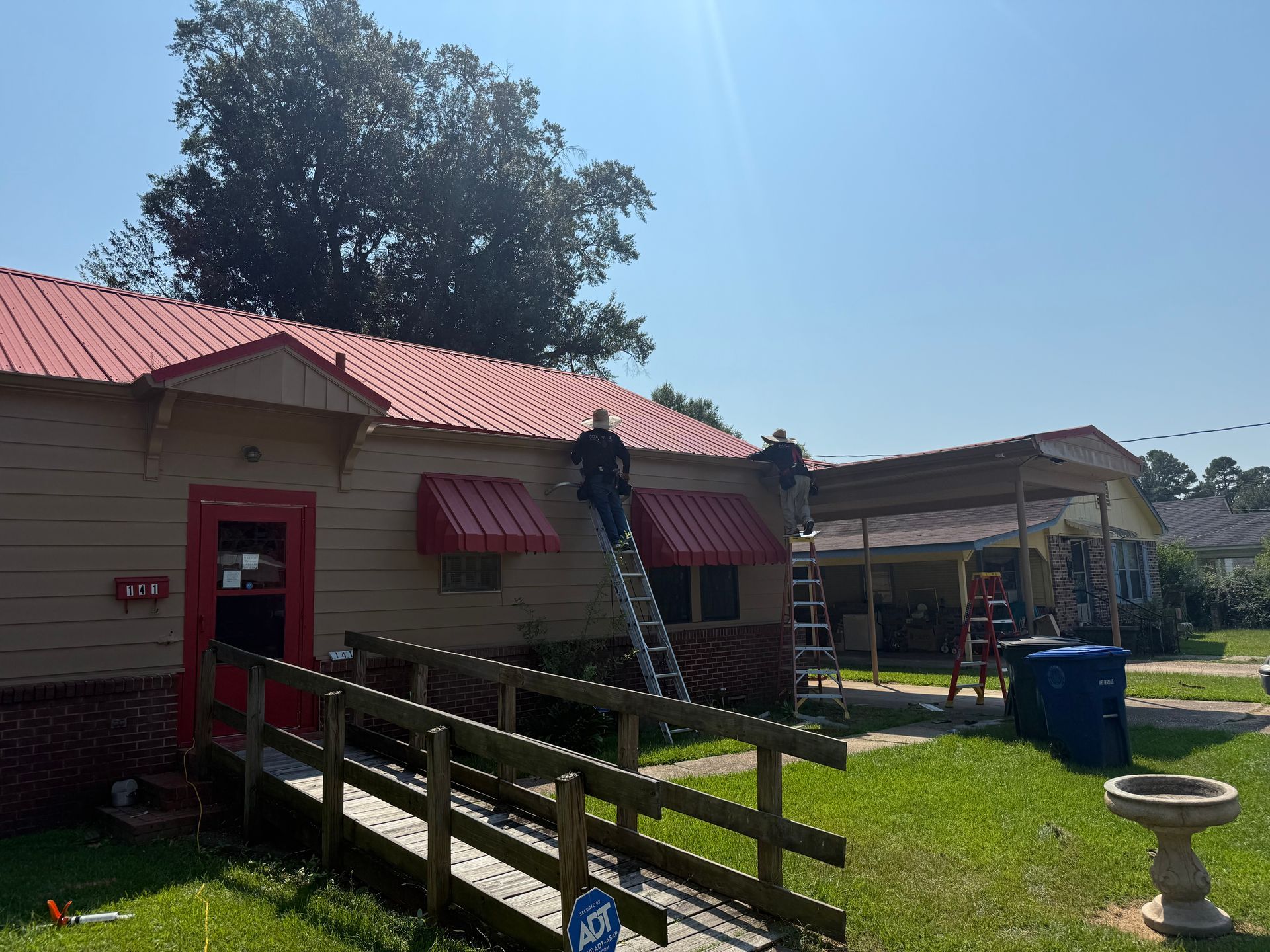 Building with a red metal roof, a ramp, and workers on ladders. Red awnings and a blue sky.