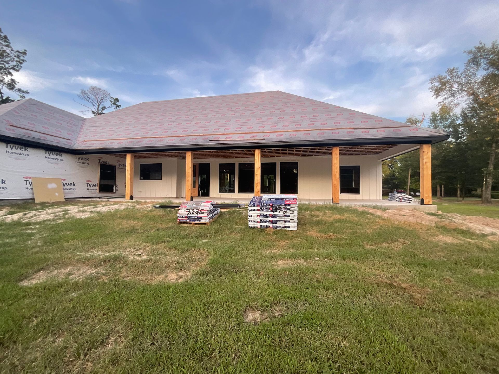 Construction of a house with a covered porch. Materials sit on the grass; the roof is being installed.