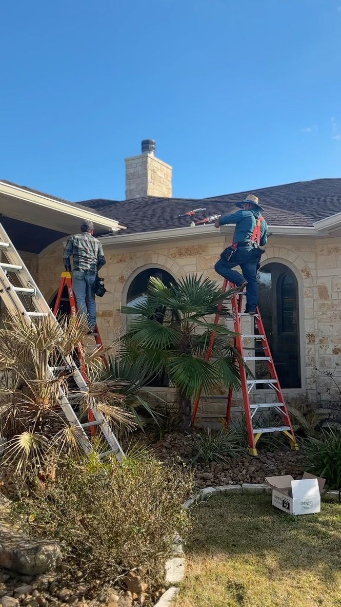Three workers on ladders installing gutters on a house with stone siding under a clear, blue sky.