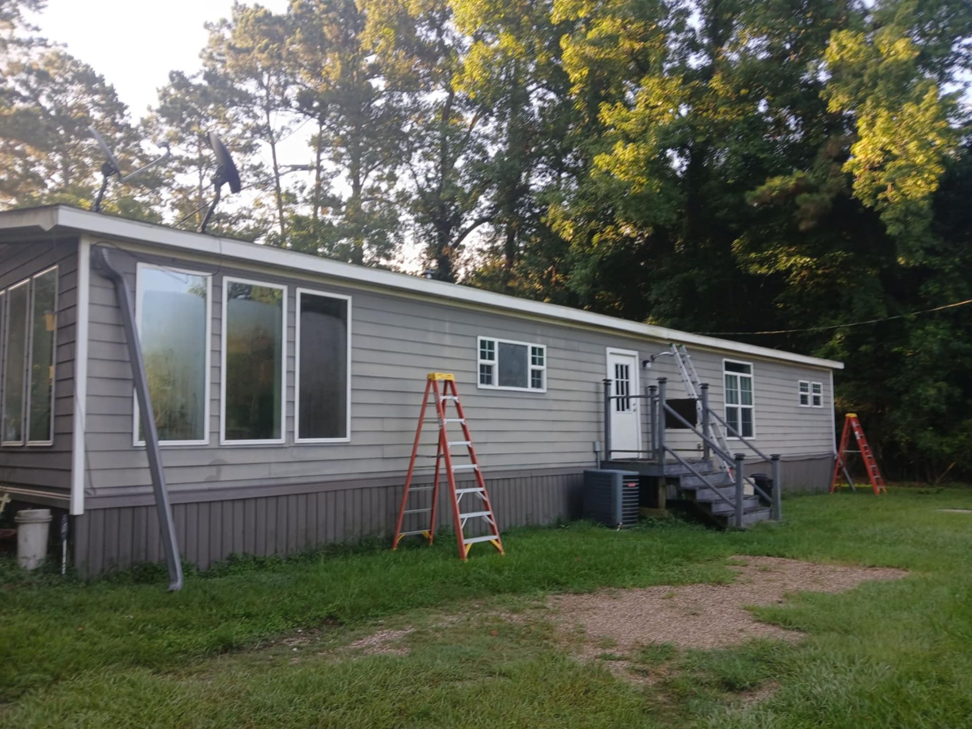 Gray mobile home with ladder, windows, and trees in a grassy yard.