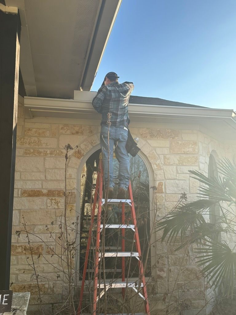 Person on ladder cleaning the gutter of a tan stone house. Sunny day.