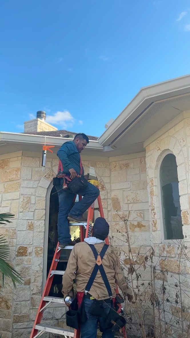 Two workers on a ladder installing gutters on a stone house under a blue sky.