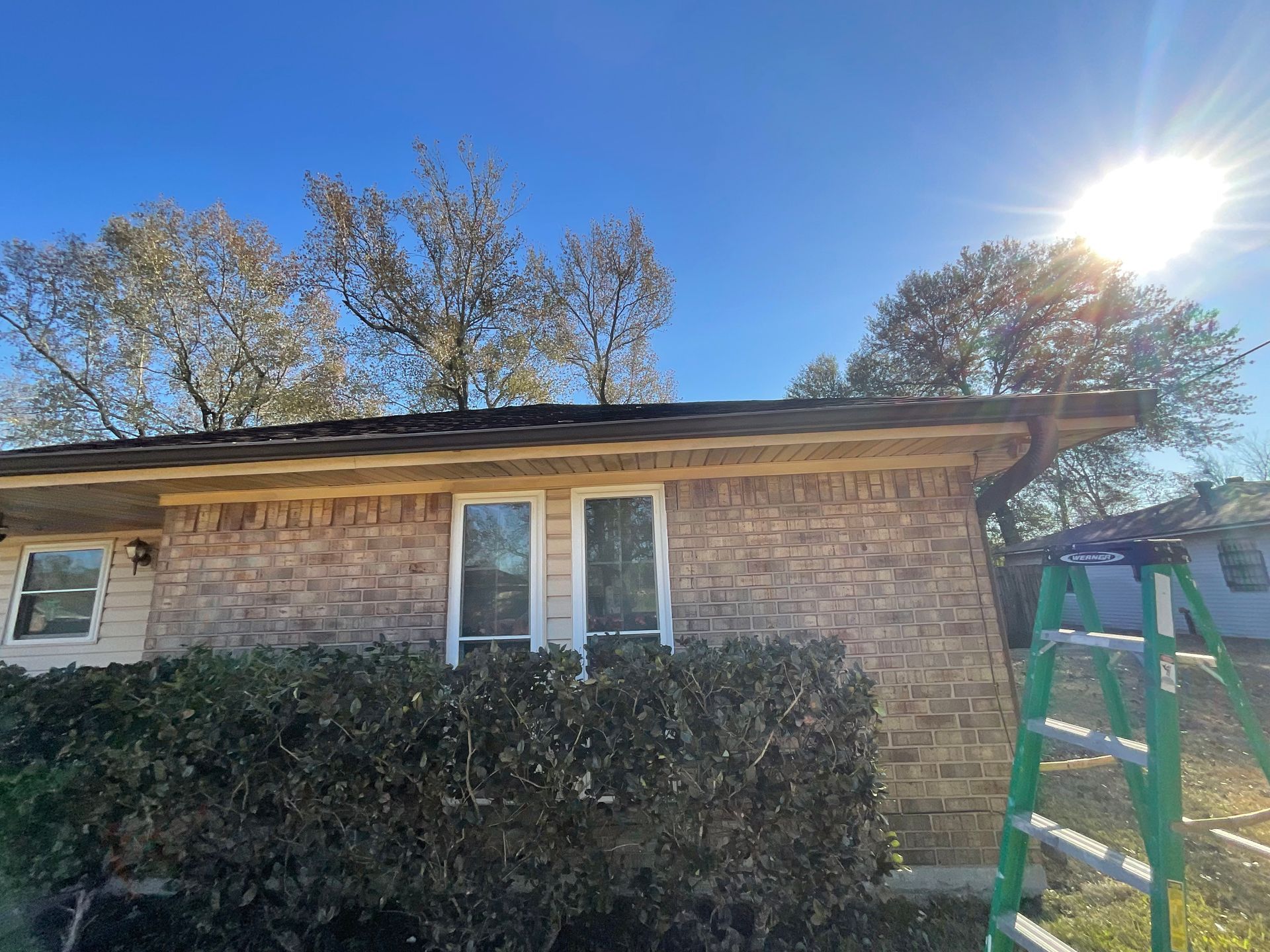 Brick house with windows, dark roof, trees, and ladder under a sunny blue sky.