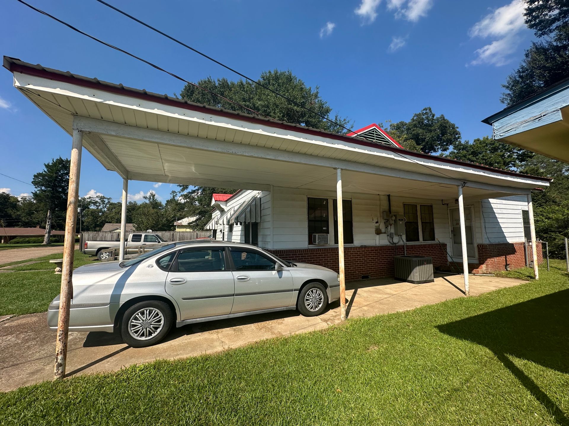 Silver car parked under a white carport attached to a white house with a red roof trim and green lawn.