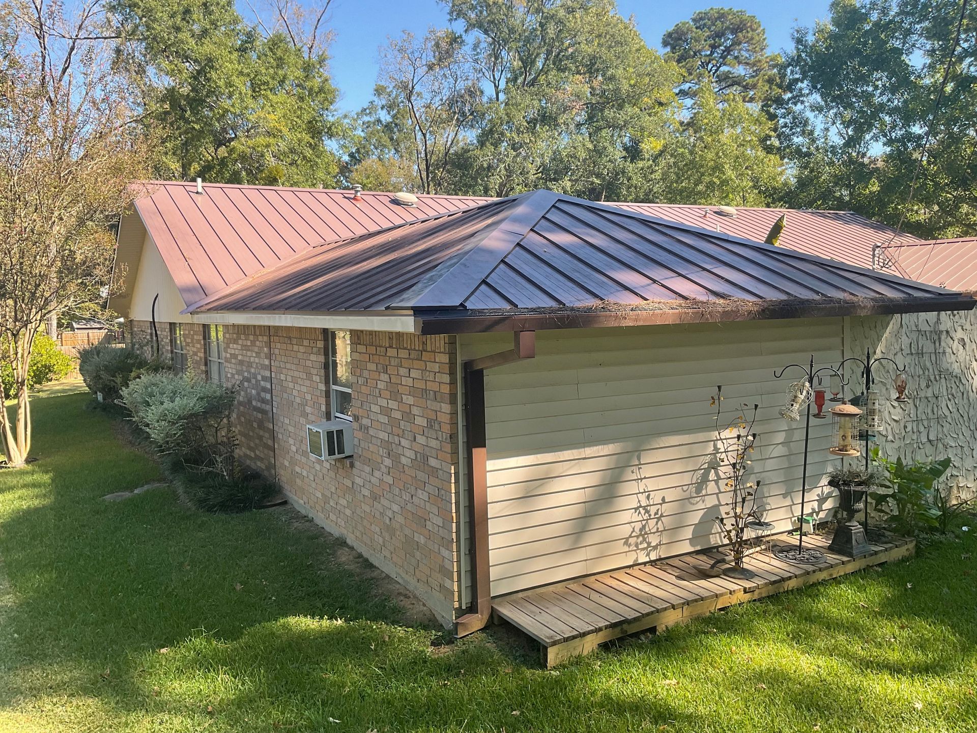 Brick house with a brown metal roof, set in a grassy yard under a blue sky.
