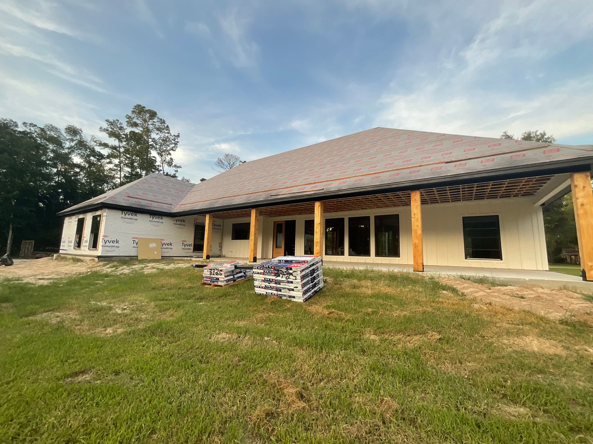 House under construction, unfinished walls, roof, and lumber supports on a grassy lot under a cloudy sky.
