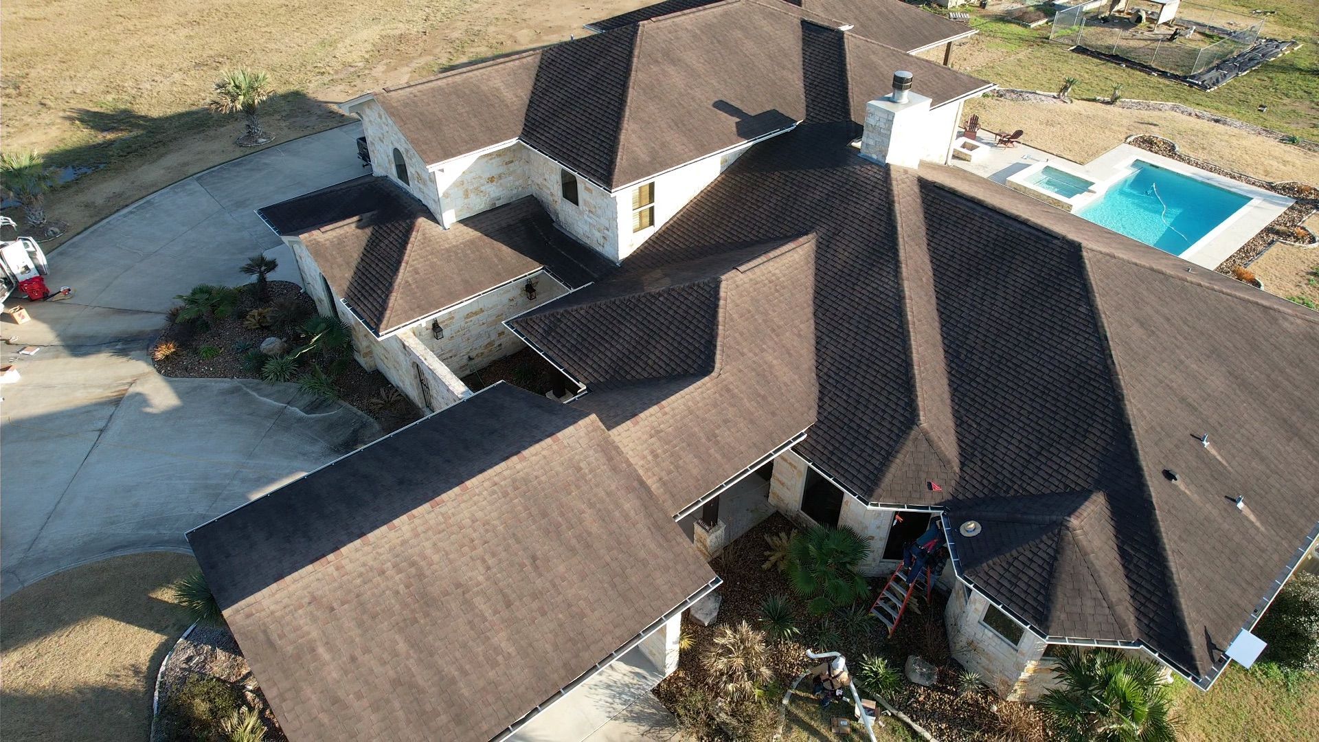 Aerial view of a large house with a dark brown roof, a pool, and a driveway in a dry, grassy area.