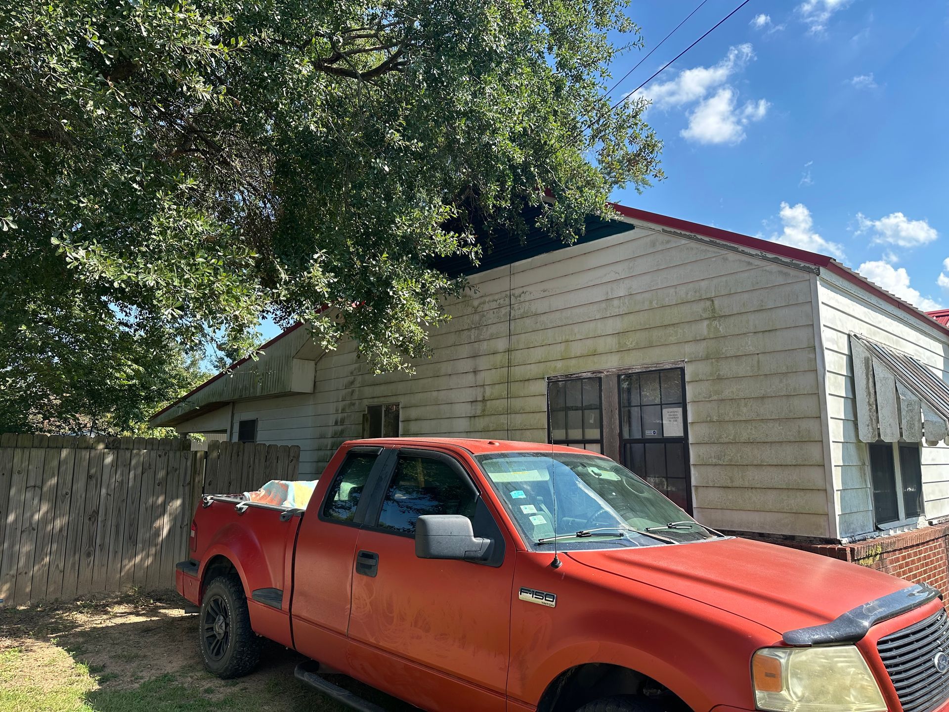 Red pickup truck parked in front of a weathered, white house with a red roof, under a tree.