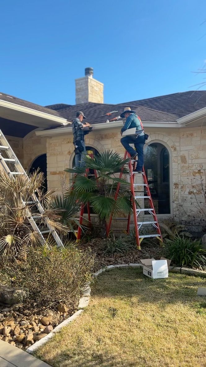 Two workers on ladders cleaning gutters of a house with stone siding on a sunny day.
