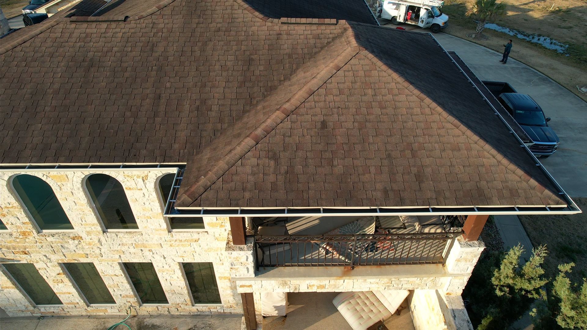Drone view of a large house with a brown roof and stone facade. A balcony and black car are visible.