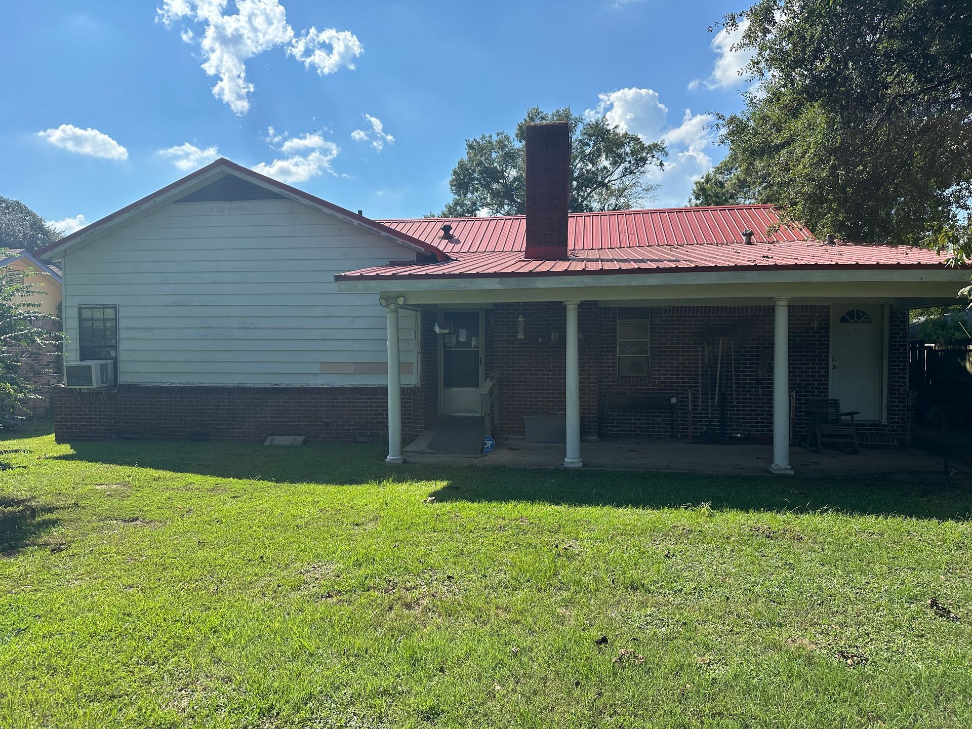 Backyard of a house with red metal roof, white siding, and covered porch with pillars.
