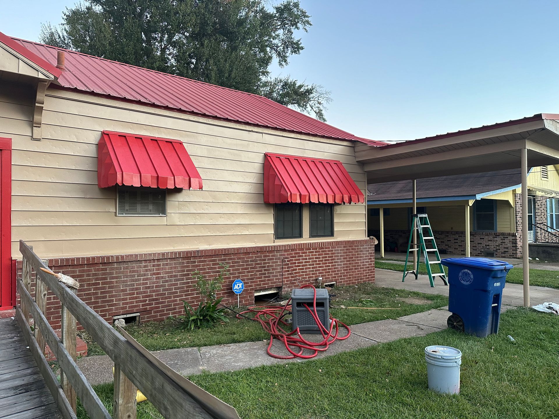 Tan house with red awnings and roof. Blue trash can and green lawn in front.